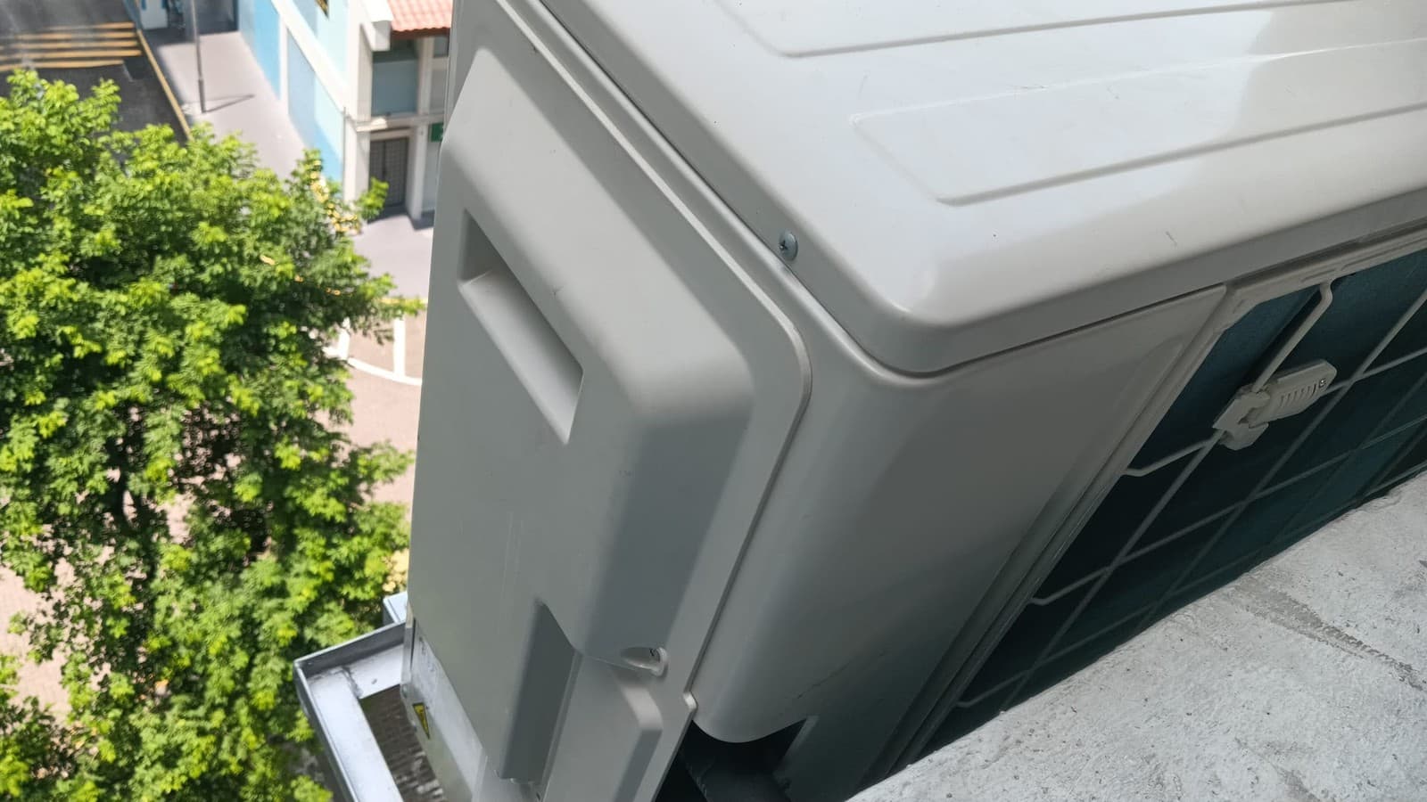 Outdoor condenser unit mounted on an aircon ledge of an HDB block, with trees visible behind it