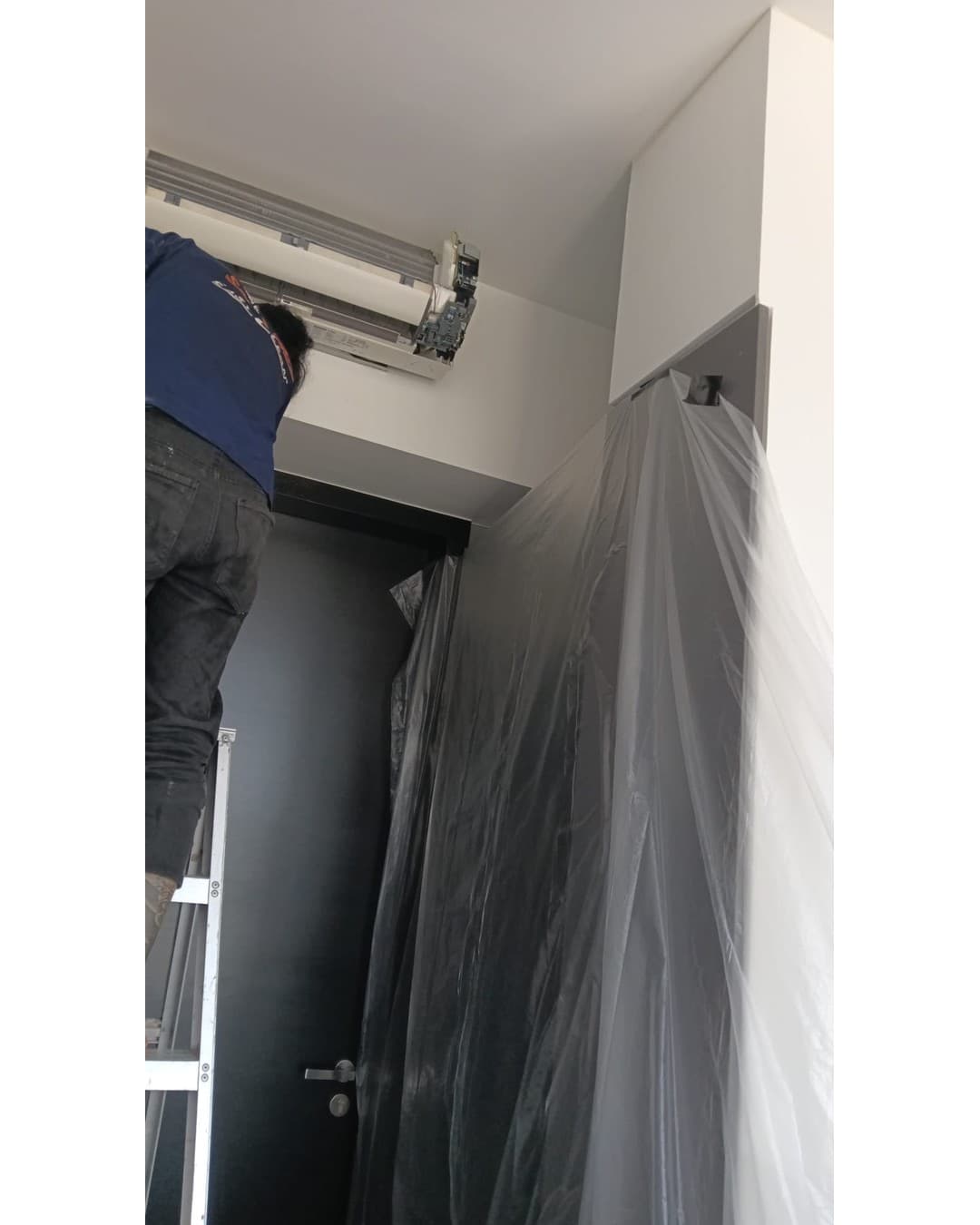 Cool Aircon technician on a step ladder working on a wall-mounted aircon unit, with plastic sheeting hung to protect the wardrobe below