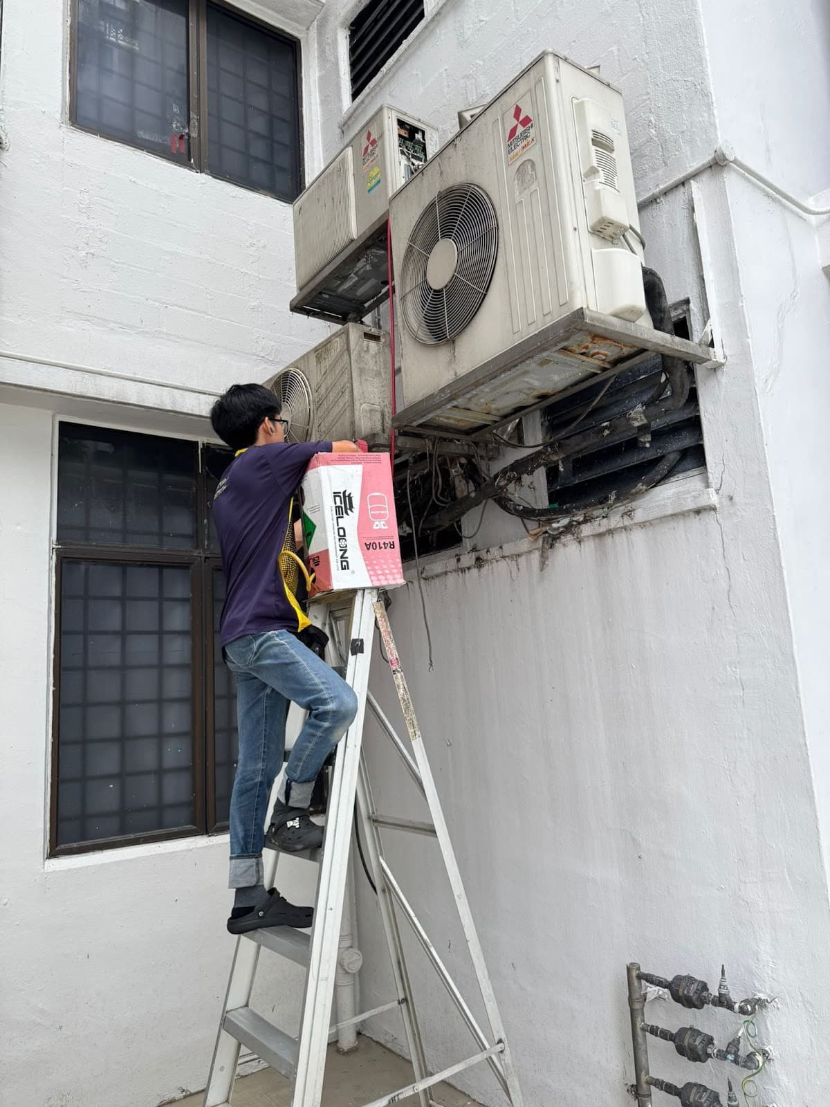 Technician on a ladder working on two outdoor aircon units mounted on an outside wall